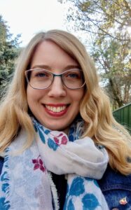 Smiling woman with blonde hair and glasses outdoors, wearing a patterned scarf and denim jacket, trees in the background.